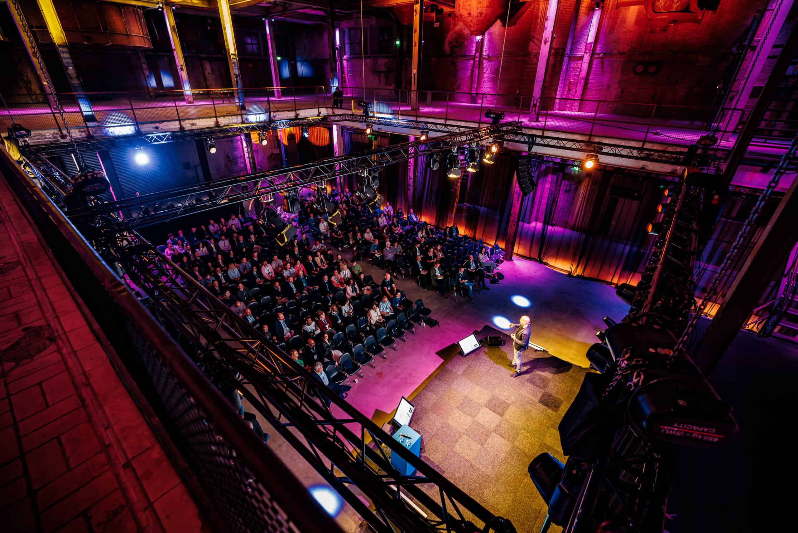 Grote zaal in de Middenfabriek met publiek en presentator tijdens evenement.