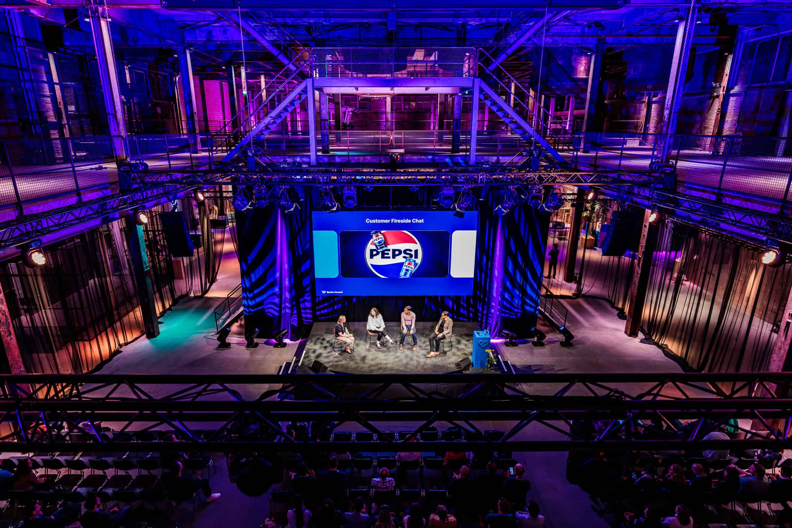 Evenement in de Middenfabriek met podium en publiek, verlicht in blauw en paars.