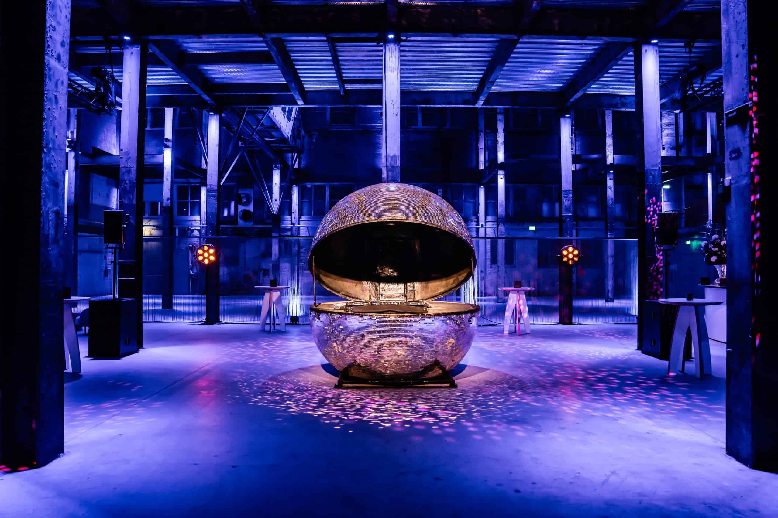 Middenfabriek met grote, ronde sculptuur in industriële setting, verlicht met blauwe en paarse licht.