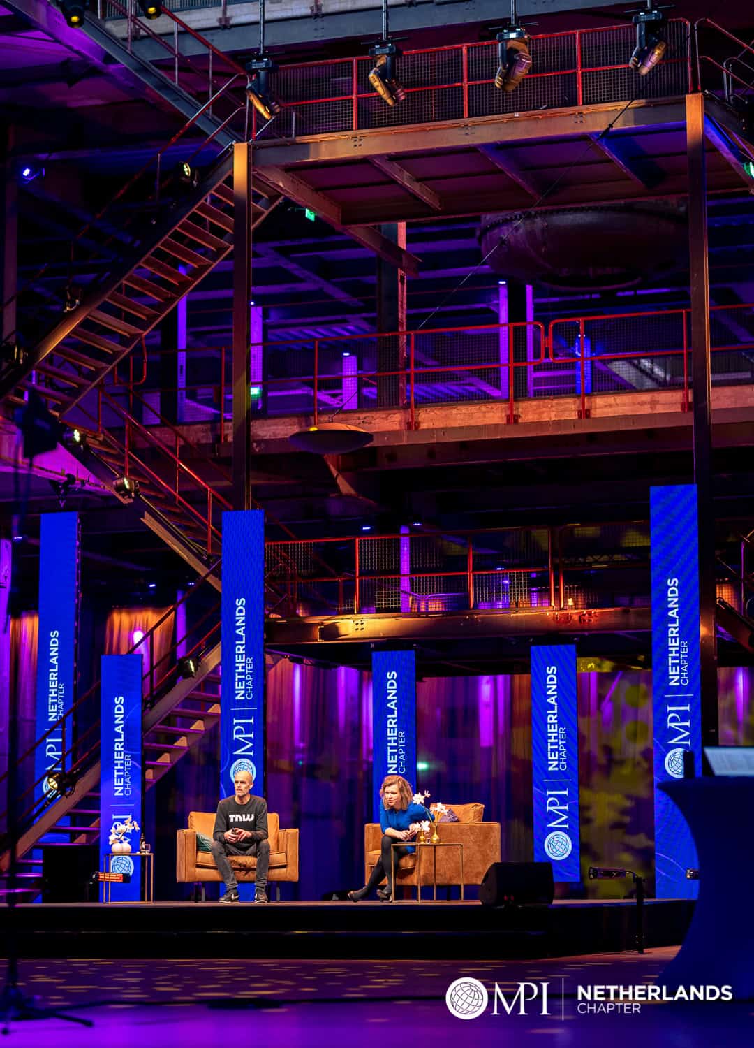 Twee sprekers op het podium tijdens MPI Online Event met kleurrijke verlichting.