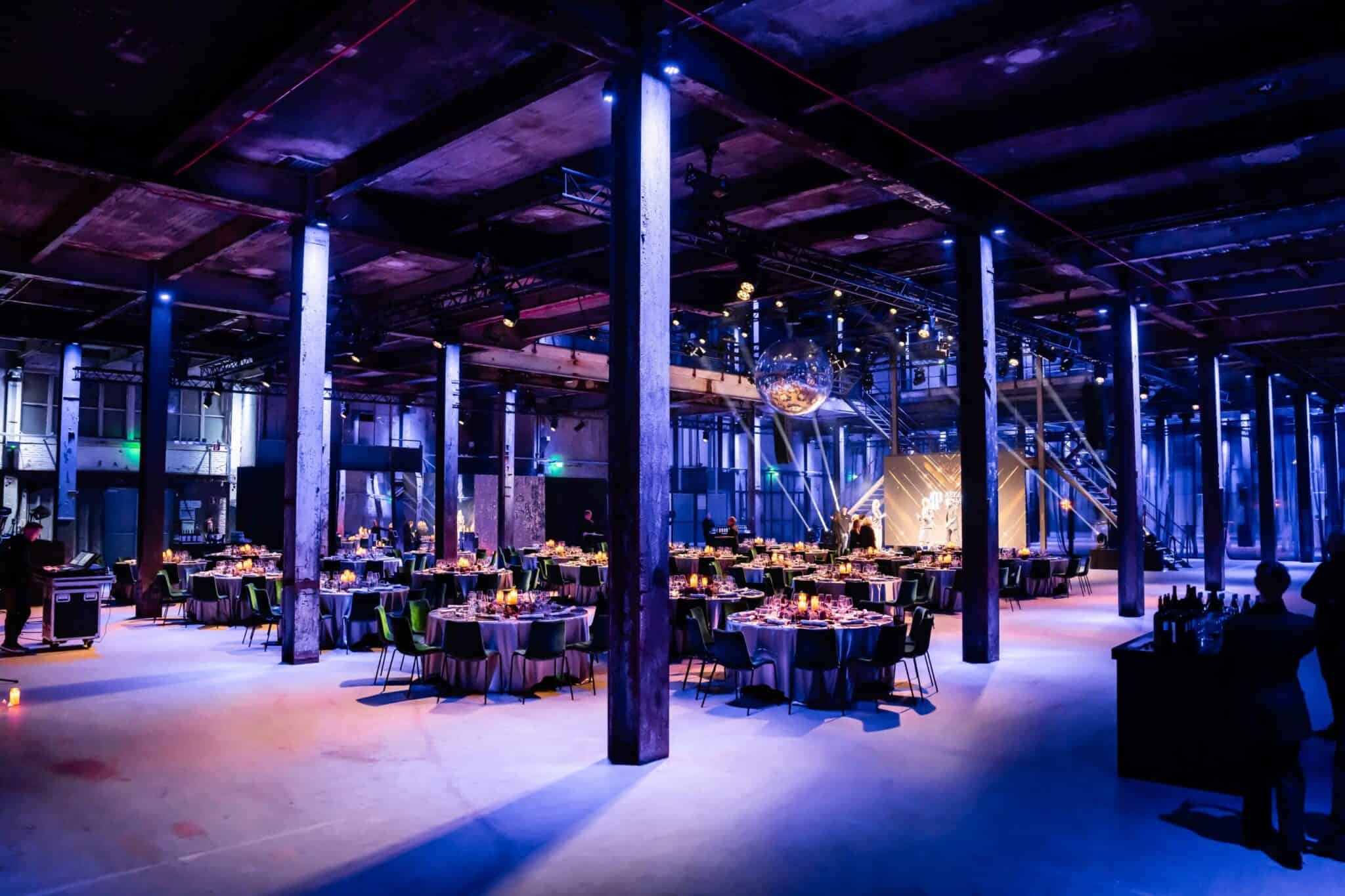 Sfeervolle awardshow in een industrieel gebouw met gedimde verlichting, ronde tafels en feestelijke.