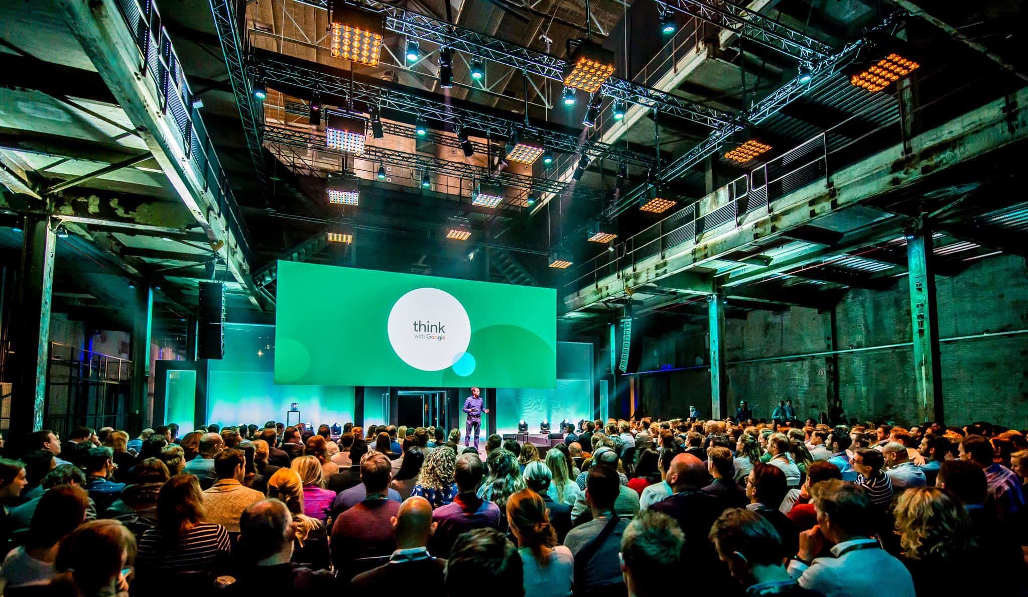 Groots congres in industriële locatie met publiek en grote schermen.