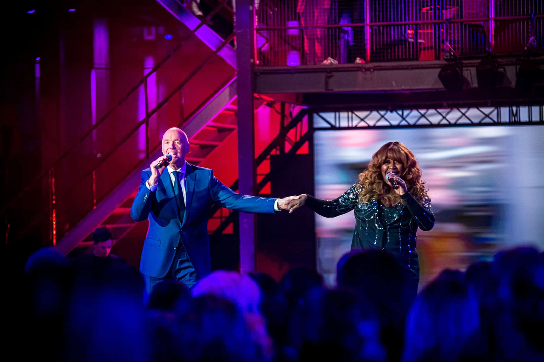 Twee artiesten zingen tijdens EO Christmas TV Show op SugarFactory, omringd door een enthousiast pub.