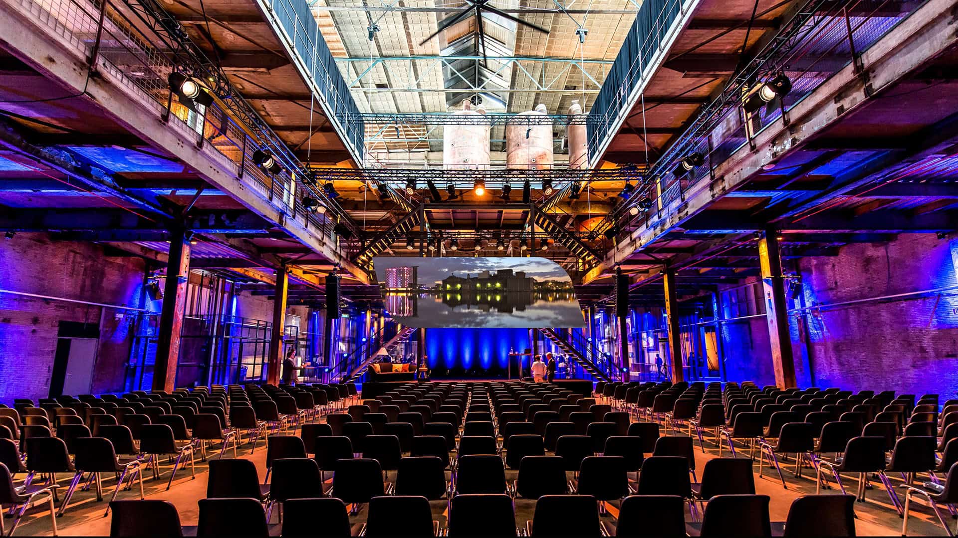 Grote industriële zaal met podium en zitplaatsen, geschikt voor evenementen en presentaties.