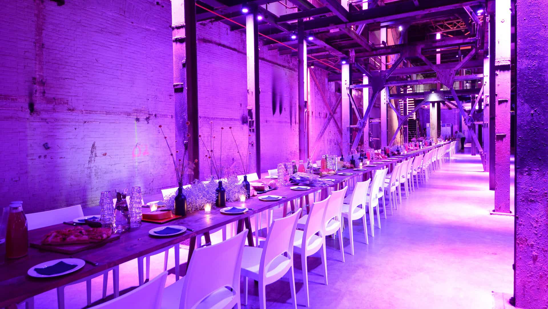 Gezellig industrieel restaurant met lange tafel onder kleurrijk LED-verlichting.