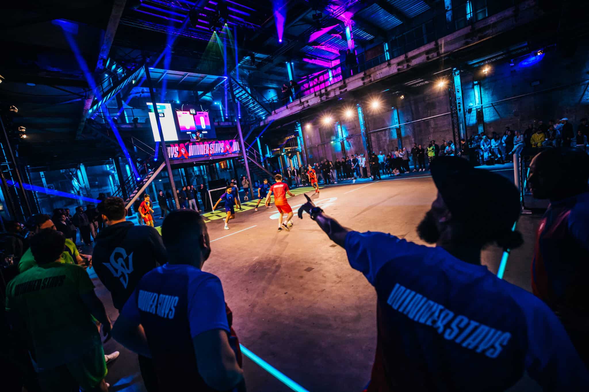 Indoor zaalvoetbalwedstrijd met spelers en publiek in een verlichte sporthal.