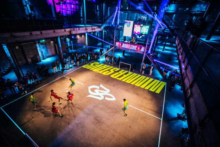 Voetbalwedstrijd in een industrieel gebouw met neonverlichting en publiek.